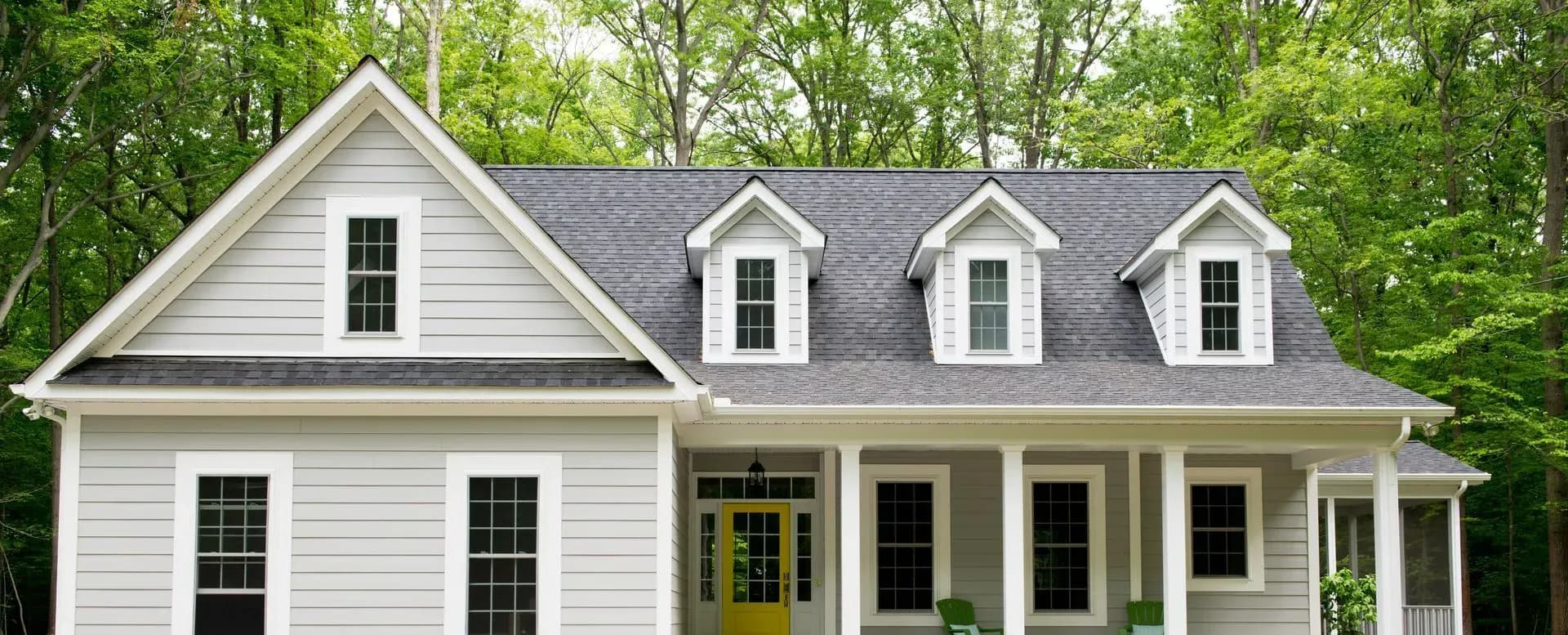A charming gray two-story house with a covered porch, three dormer windows, and a vibrant yellow front door. Set against a backdrop of lush green trees.