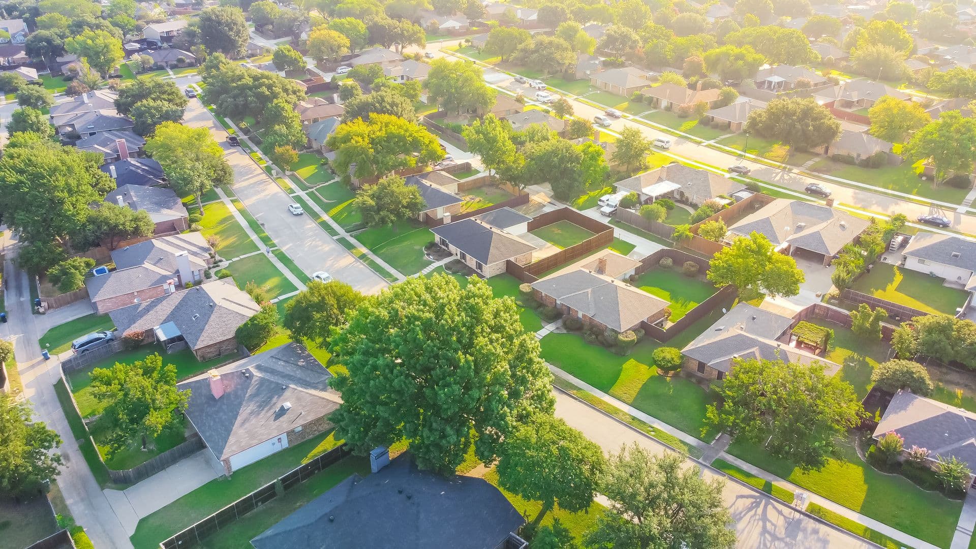 Aerial view of a suburban neighborhood with tree-lined streets and single-family homes. Bright, sunny day with lush green lawns and a peaceful atmosphere.