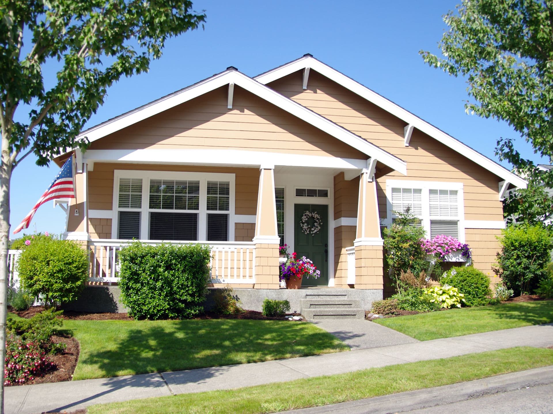 Charming tan bungalow with white trim, surrounded by lush landscaping and colorful flowers. An American flag waves beside the porch. Bright, sunny day.