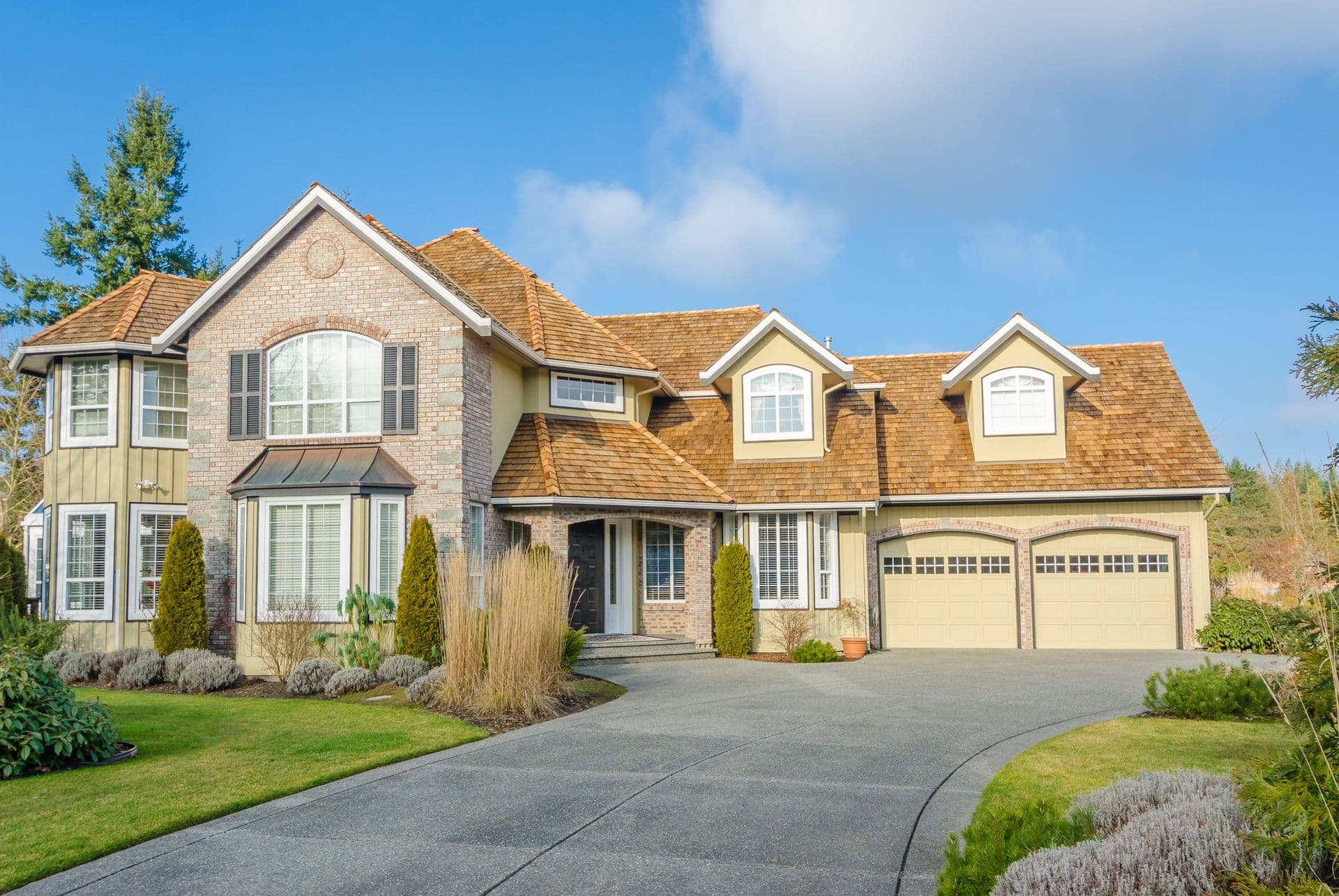 A large, elegant suburban house with brick and siding exterior, featuring multiple gables and windows. It's surrounded by a manicured lawn and shrubs under a clear blue sky.