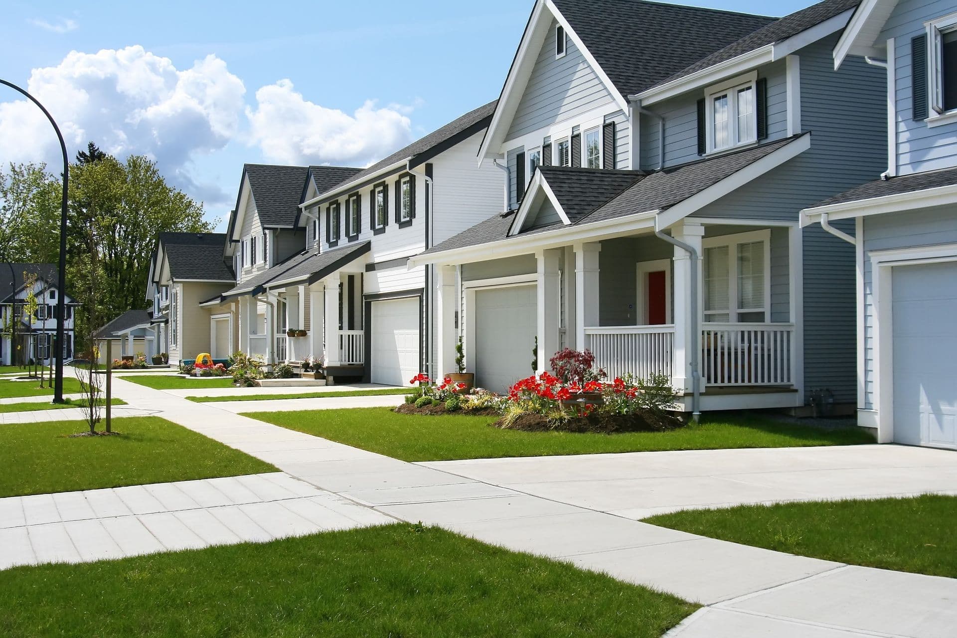 A row of modern suburban houses with white siding and dark roofs. The homes have neat lawns, flower beds, and a bright, clear blue sky above.