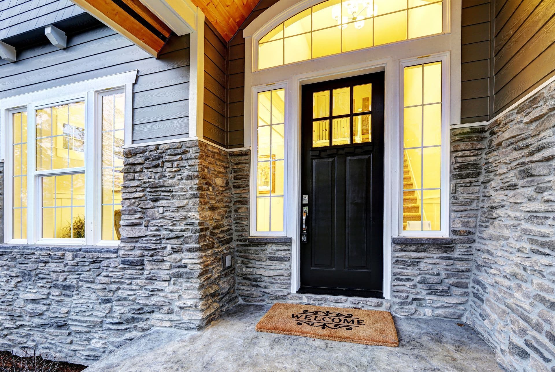 Front entrance of a modern home with a black door framed by tall windows and stone accents. A welcome mat creates a warm, inviting atmosphere.
