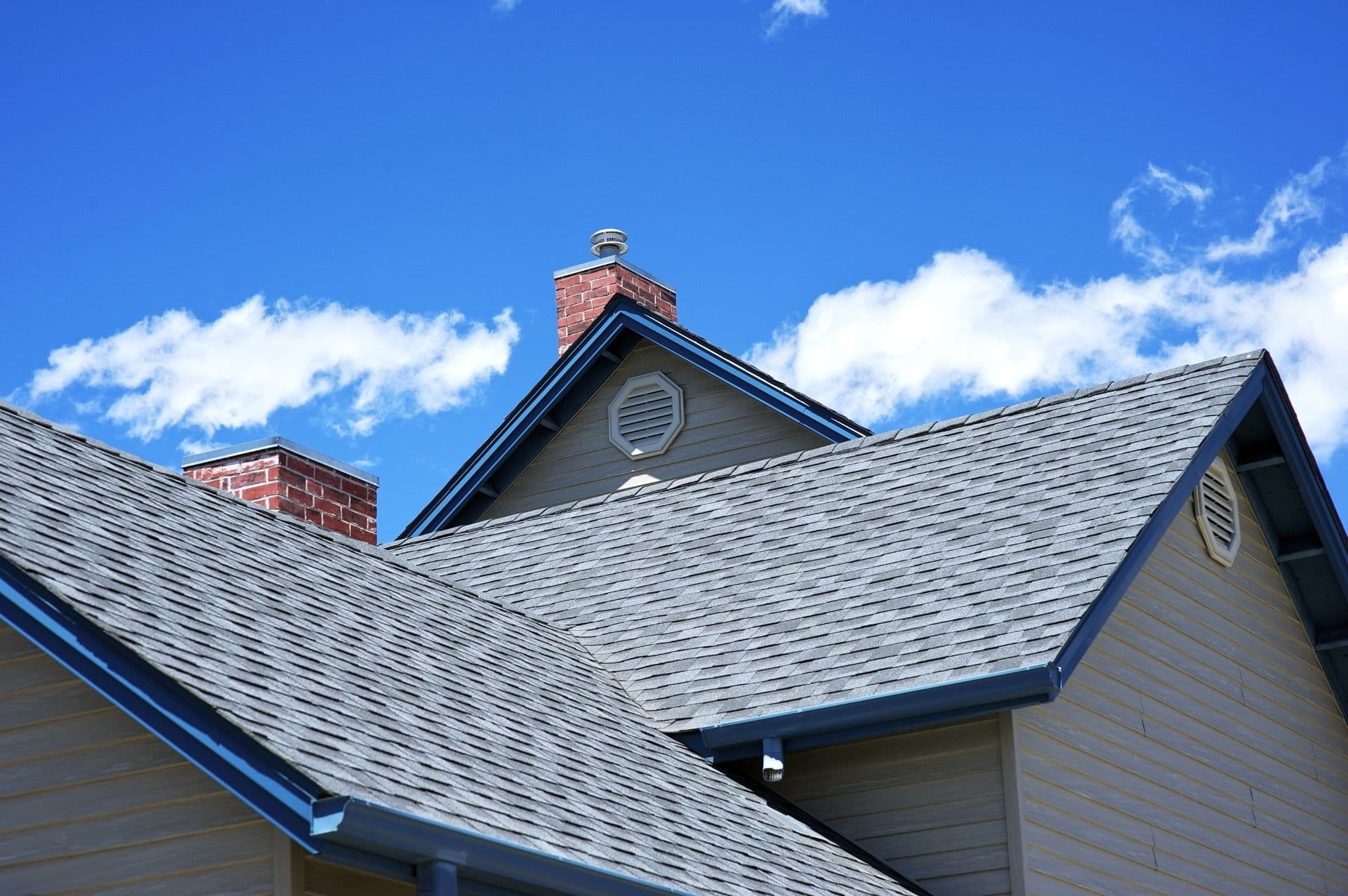 Gabled roof with gray shingles against a vibrant blue sky, featuring red brick chimneys and decorative attic vents, conveying a serene, sunny day.