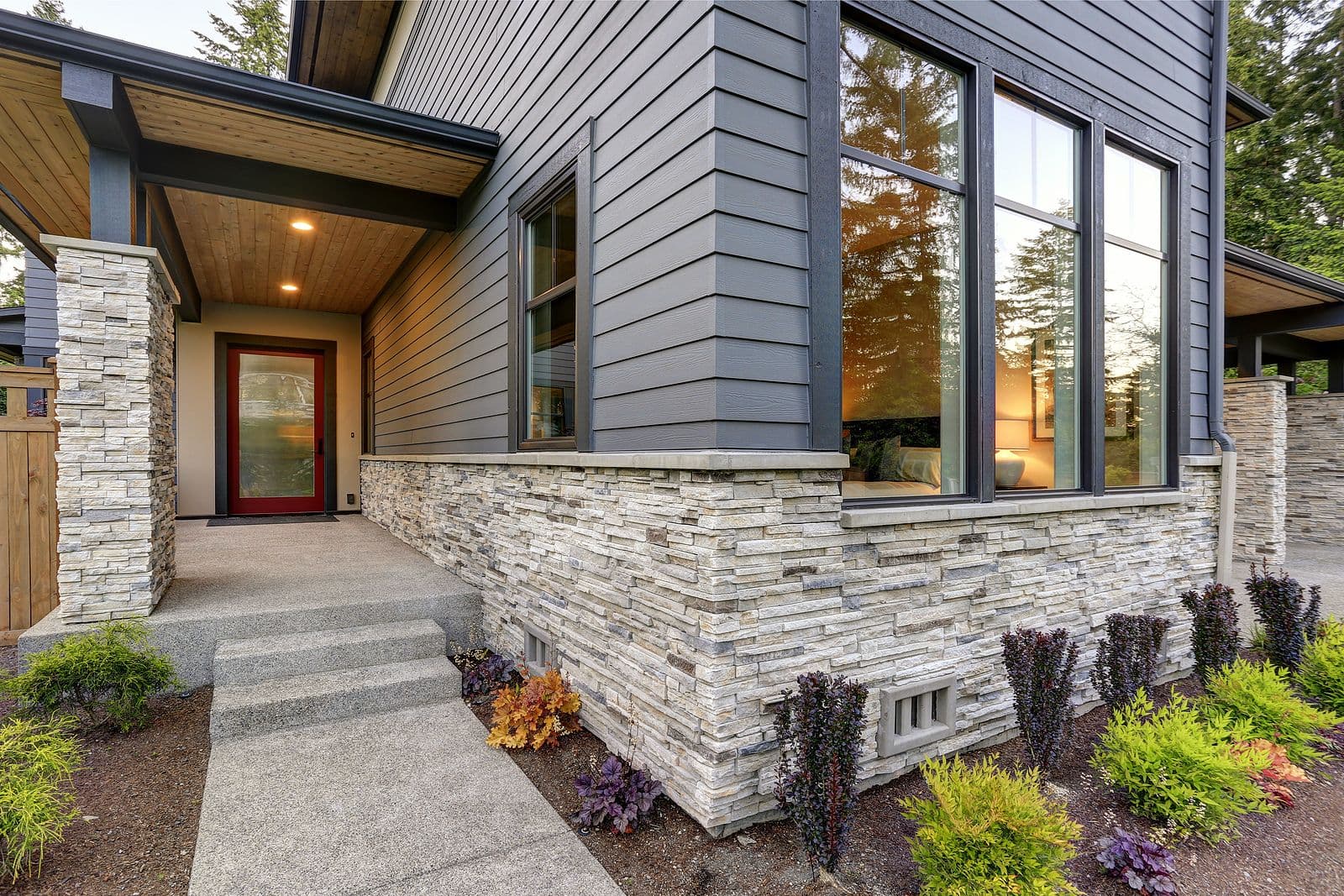 Modern home's front exterior with gray siding, stone accent walls, large windows, and landscaped path. Warm interior light visible inside.