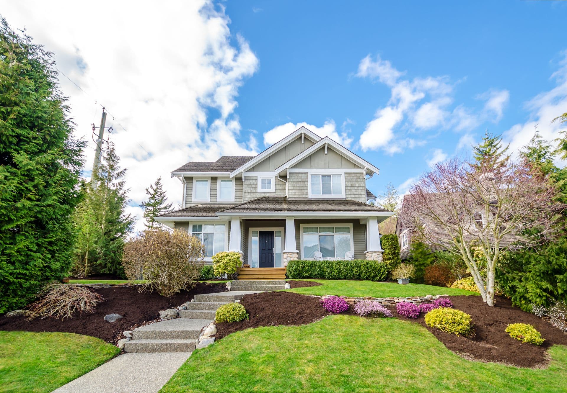 A two-story gray house with gabled roof, surrounded by manicured shrubs and colorful flower beds, under a bright blue sky dotted with clouds.