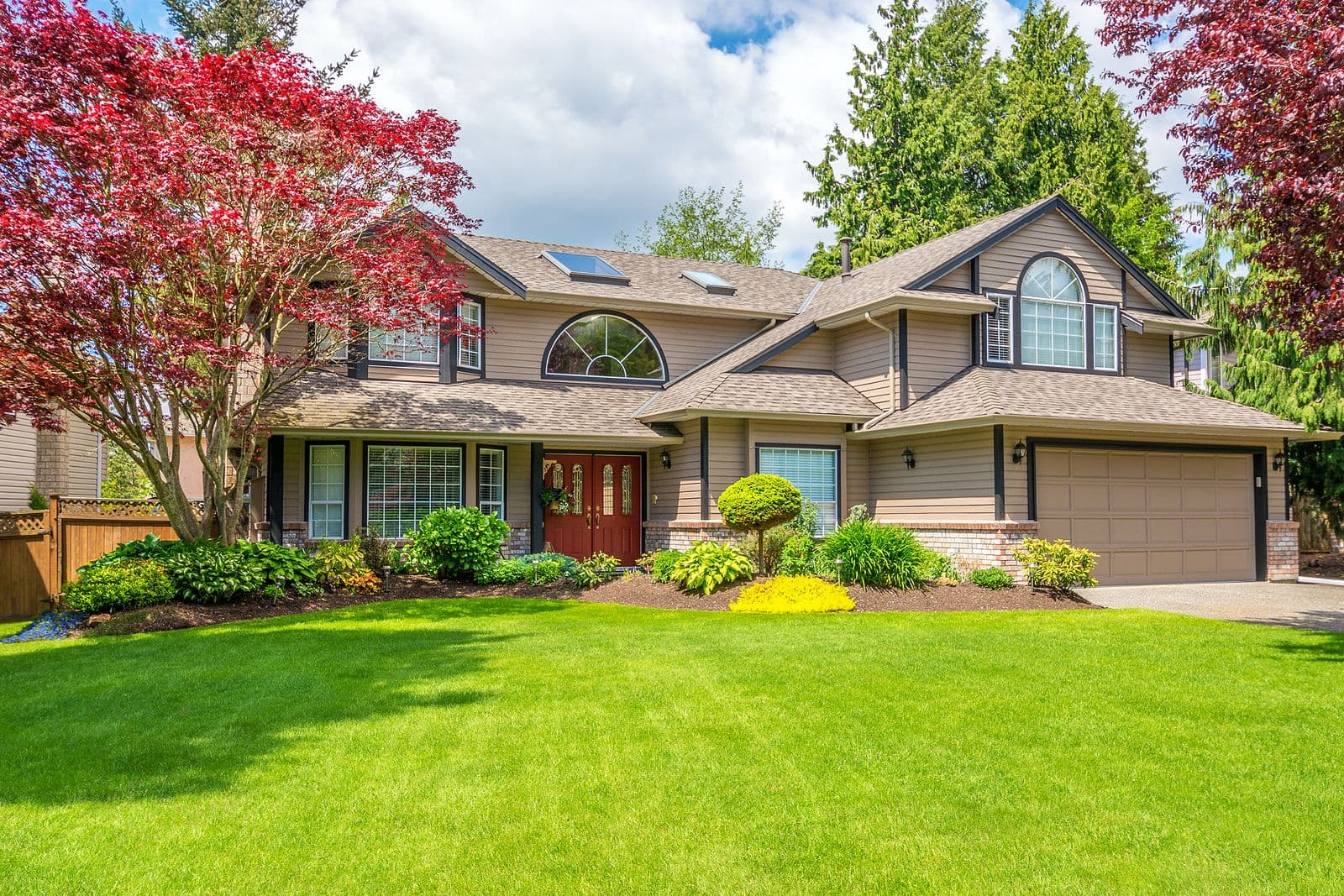 Large suburban house with a manicured lawn, vibrant red maple trees, and bushes. The home has a two-car garage, stone details, and a welcoming entrance.