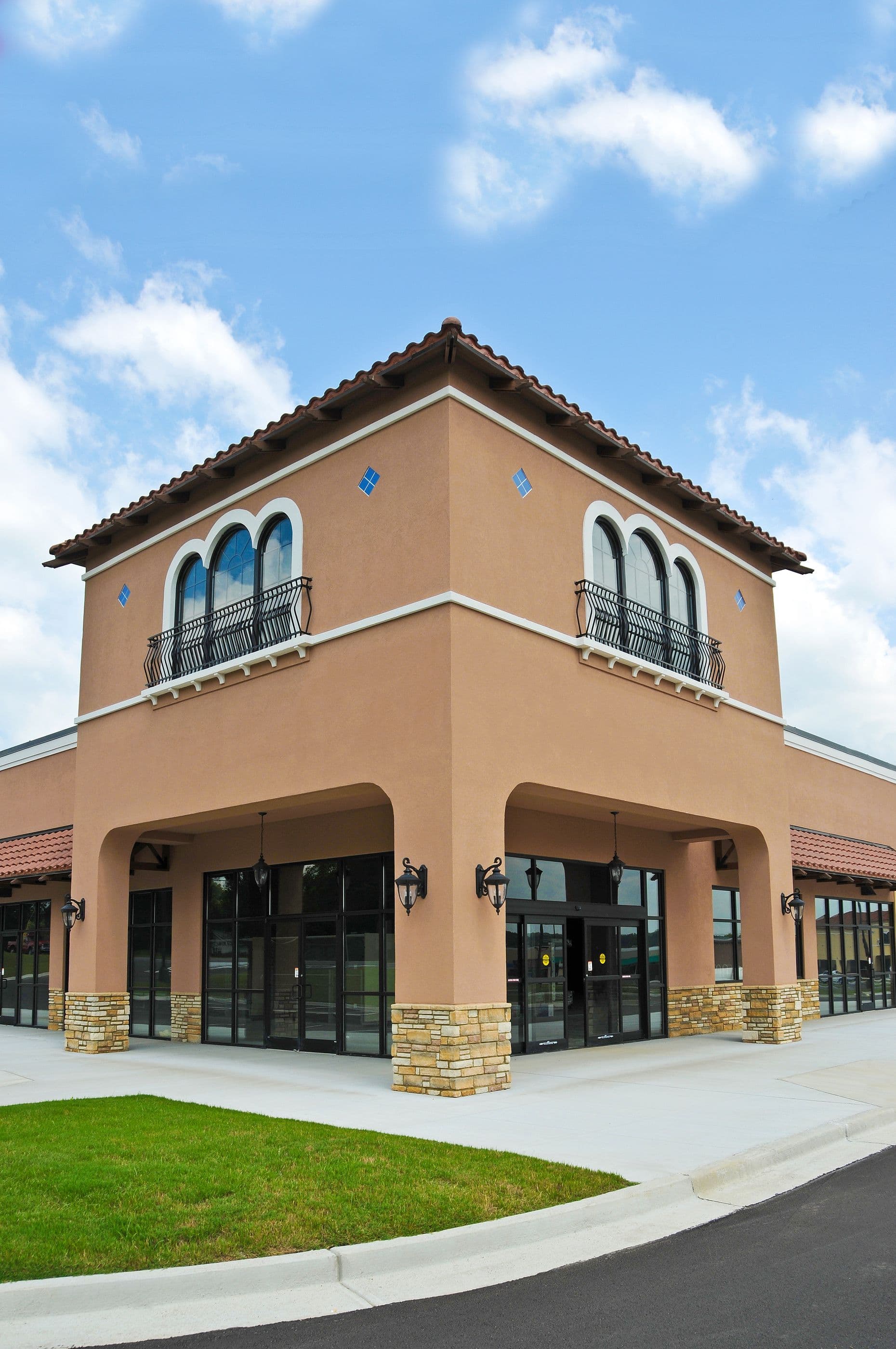 A two-story, Mediterranean-style building with arched windows and wrought iron balconies, featuring stone accents, sits under a clear blue sky.