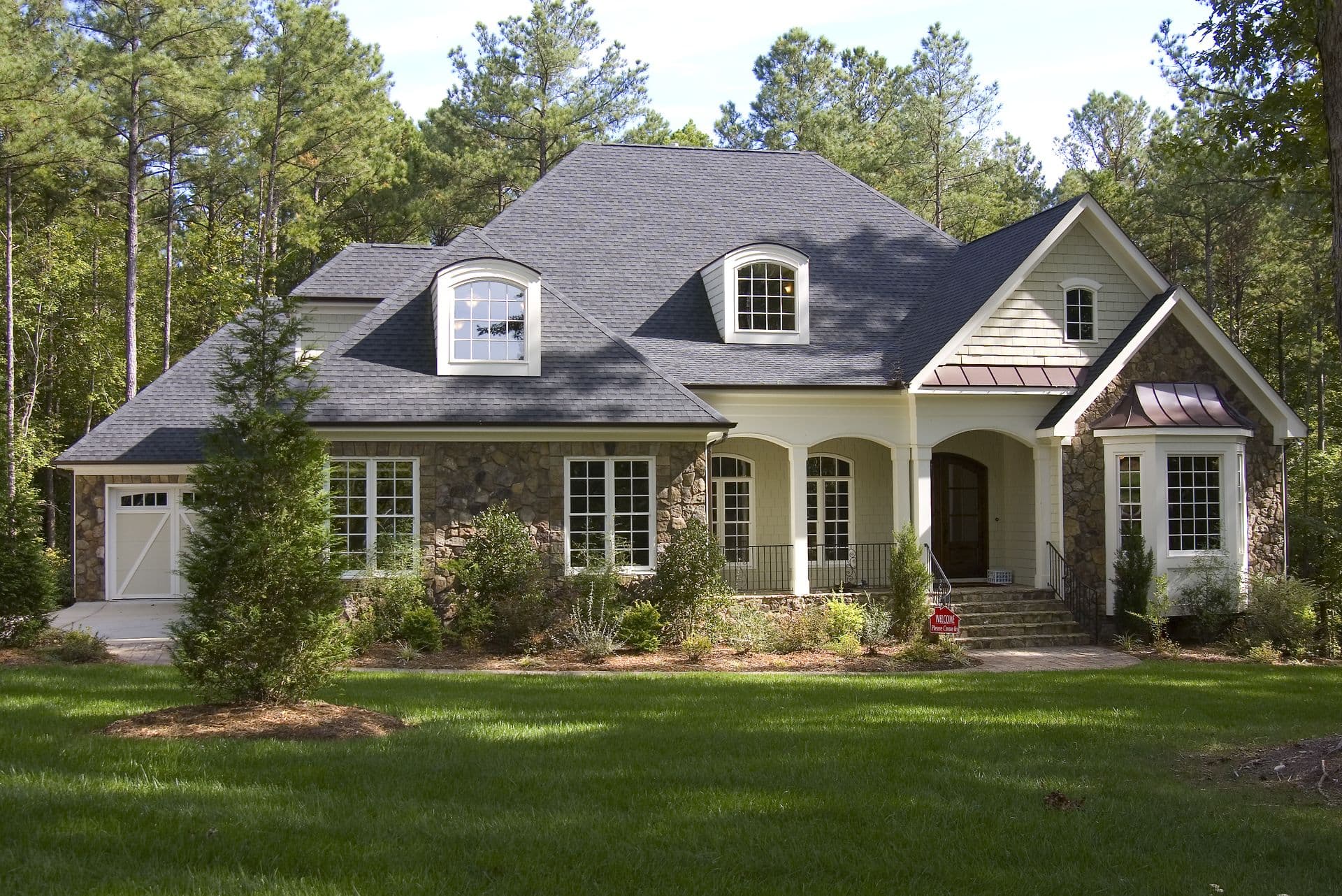 Charming suburban house with a stone facade, dark gray roof, arched windows, and lush green lawn, surrounded by tall pine trees under a clear blue sky.