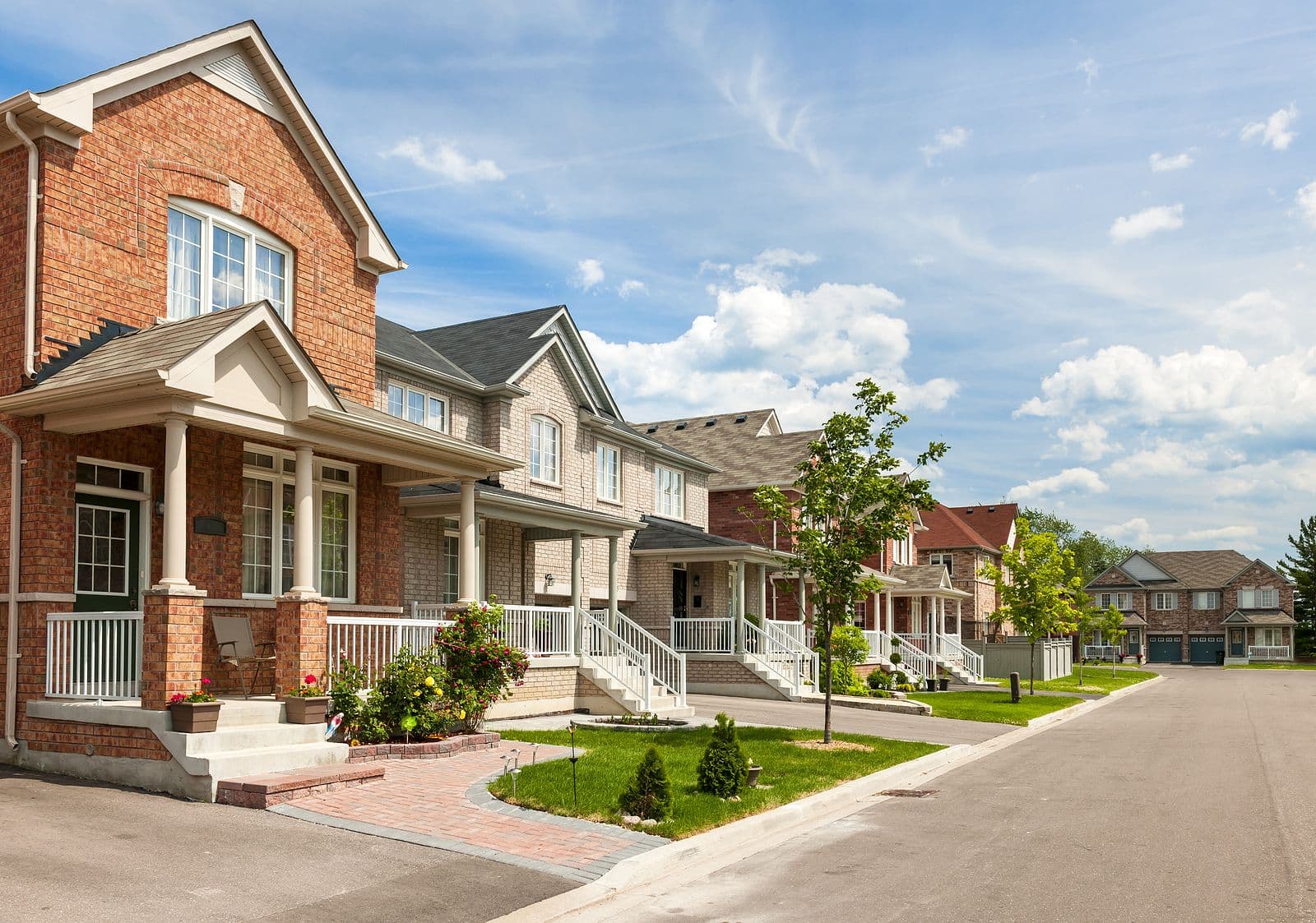 Suburban street lined with modern two-story brick houses, neatly manicured lawns, young trees, and a bright blue sky with fluffy clouds. Peaceful and inviting.