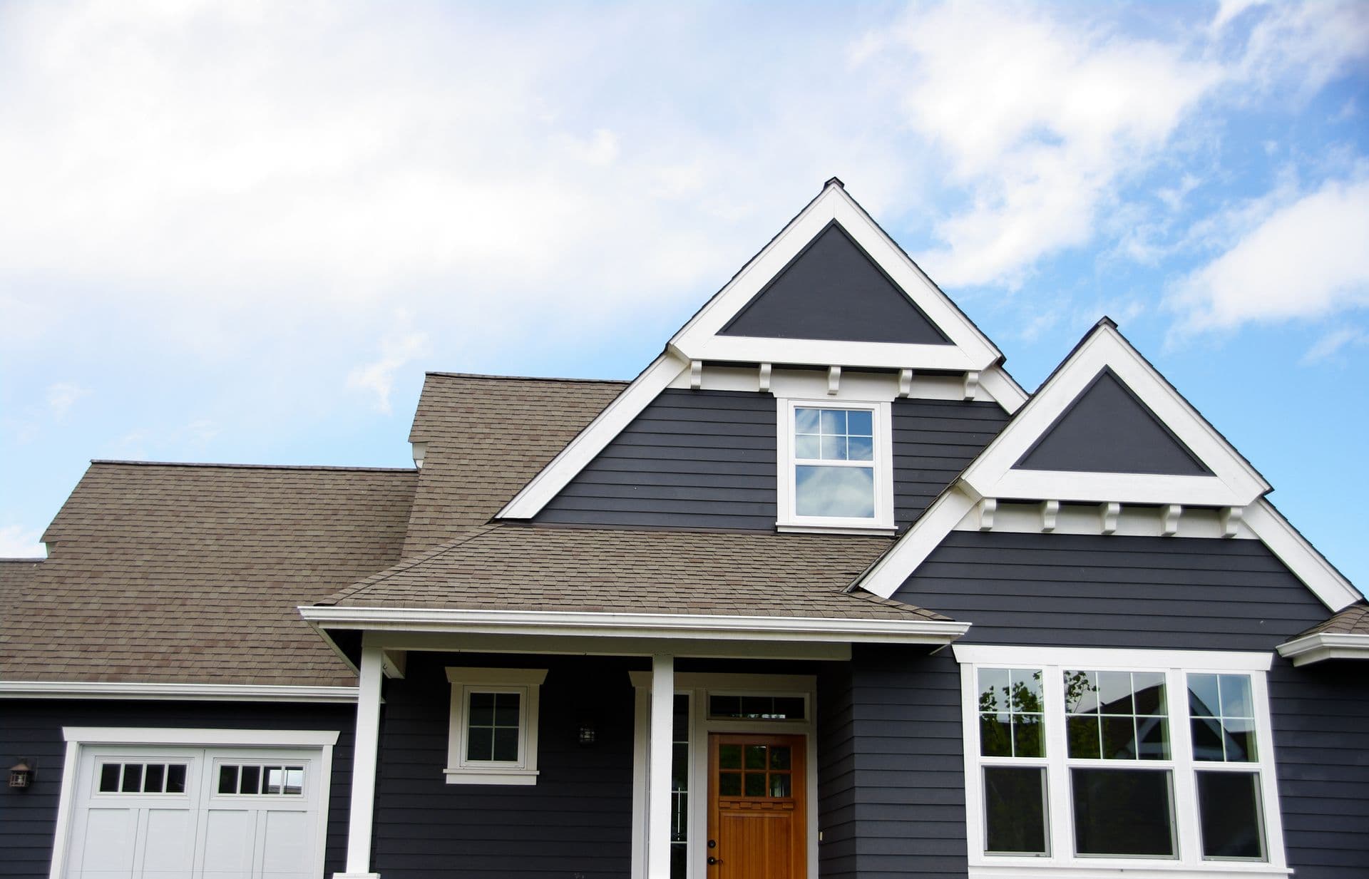 Modern suburban house with dark gray siding, white trim, and a wooden front door. Gable roof with a clear blue sky and a few clouds above.