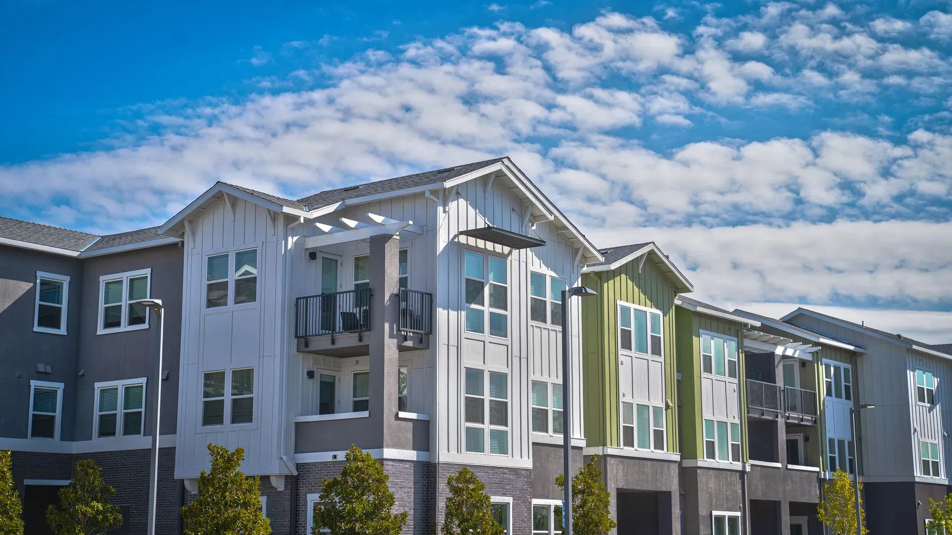Modern apartment building with varied green and gray facades under a bright blue sky with fluffy clouds. Reflective windows and small balconies add charm.