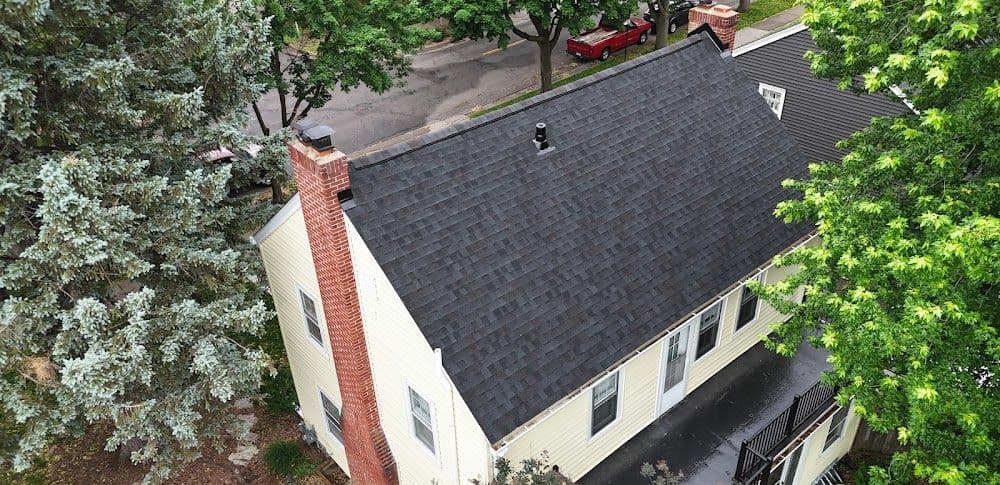 Aerial view of a house with a dark gray shingled roof and brick chimney, surrounded by lush green trees. A red car is parked on a nearby street.